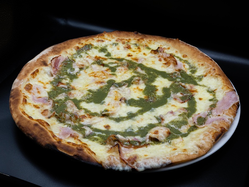 Pizza Da Kociss con mozzarella filante, prosciutto cotto e pesto verde a spirale su piatto bianco, sfondo scuro