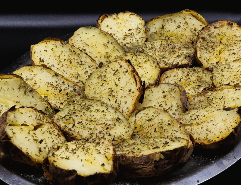 Patate al forno tagliate a metà con buccia scura e origano secco, disposte su un vassoio di metallo su fondo nero